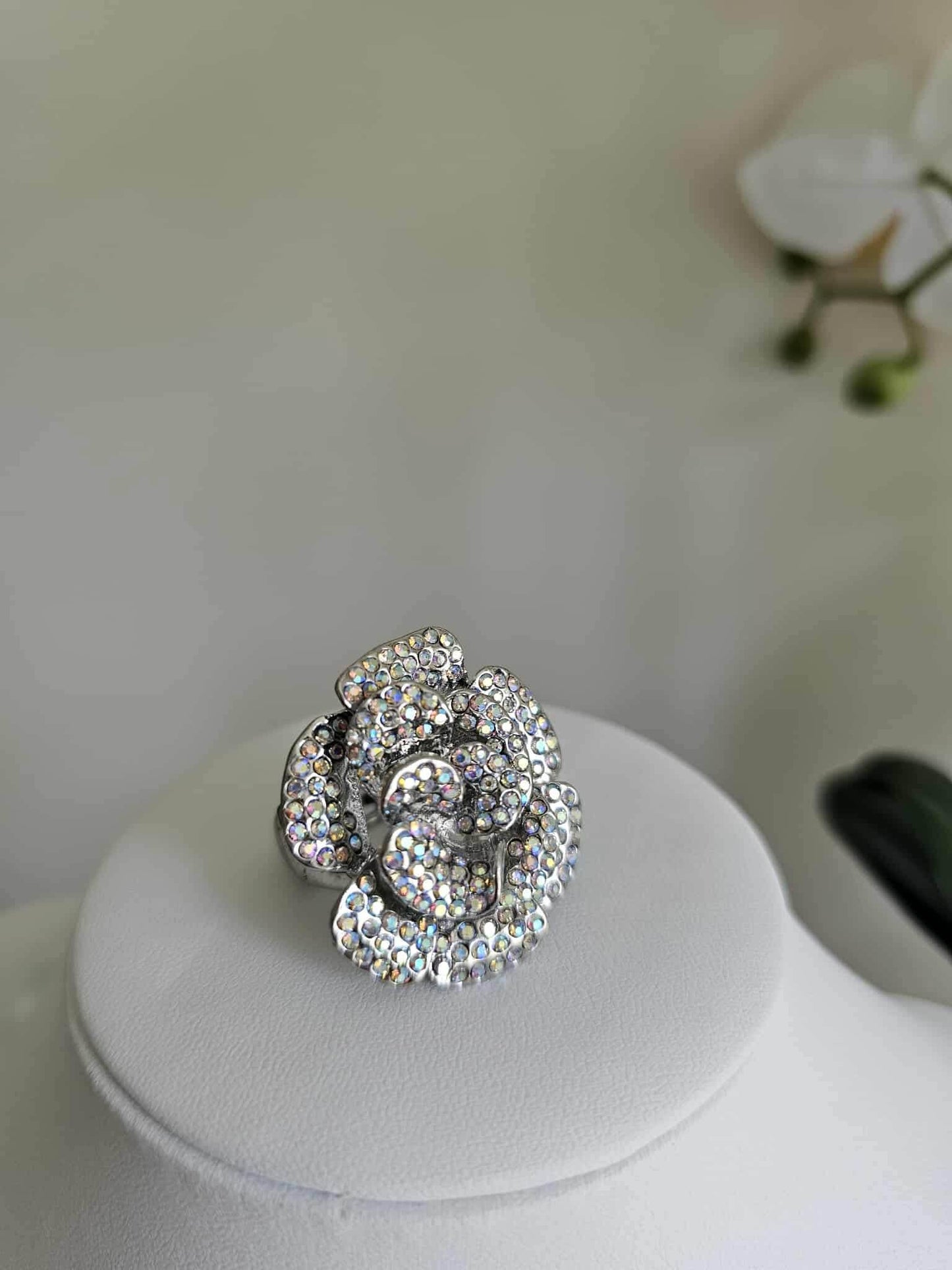 The Glamour Bloom - Silver Ring, adorned with sparkling rhinestones in a floral design, makes a dazzling statement displayed on a white round stand with a softly blurred background.