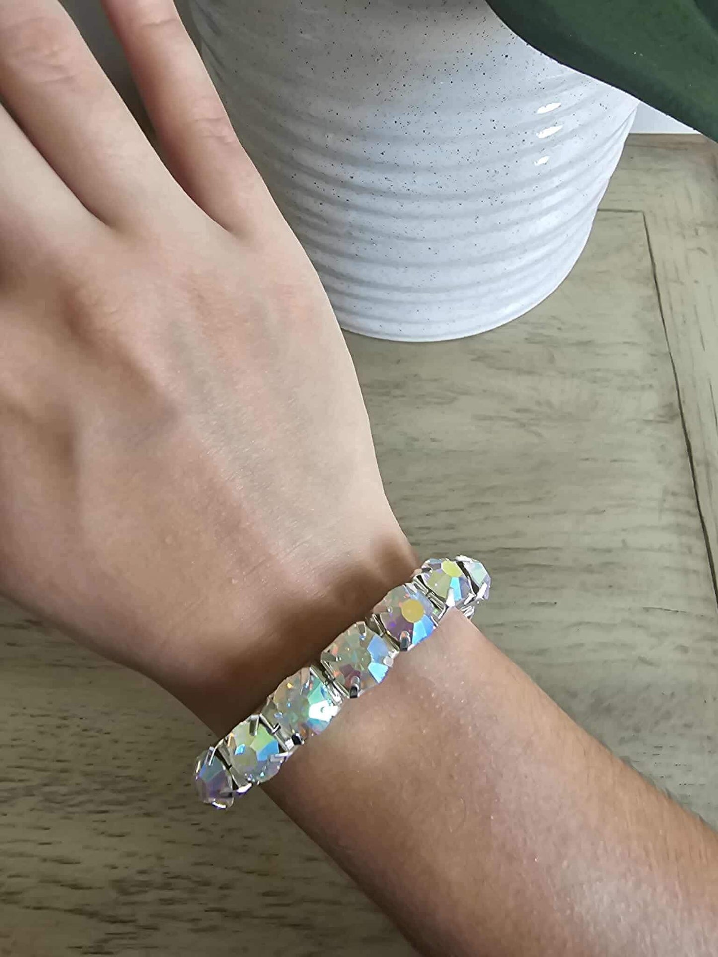 A hand wearing the Electric Elegance - Iridescent Bracelet, featuring large iridescent stones, rests on a wooden surface beside a white textured plant pot with a green leaf.