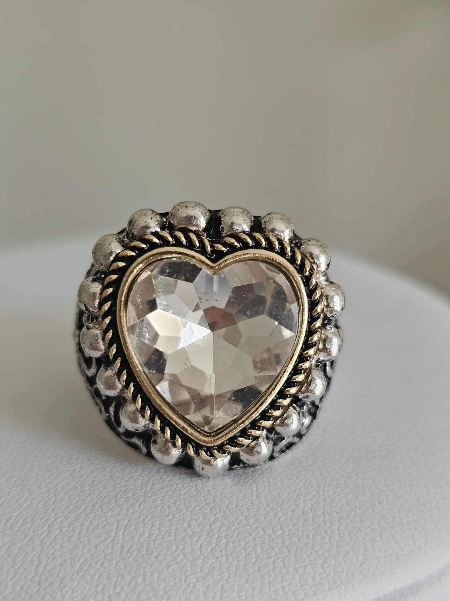 The Queen of Hearts - Silver Ring features a large, faceted heart-shaped crystal at its center, accented by a gold beaded border and displayed on a white surface.