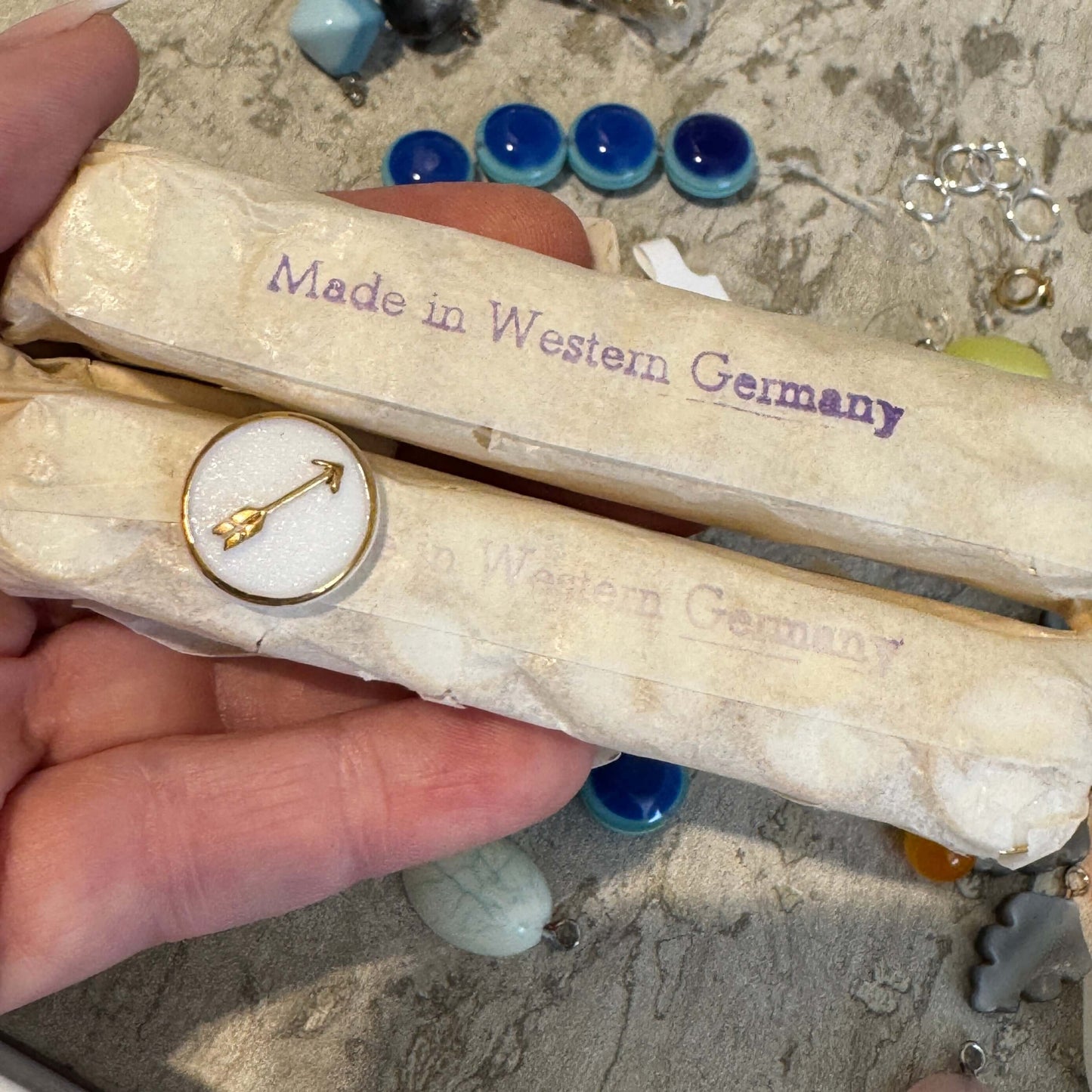 A hand holds two vintage packages labeled "Made in Western Germany" in purple. One features the Gold & White Directions Stud Earrings by Leetie Lovendale. Colorful beads and jewelry findings are scattered in the background.