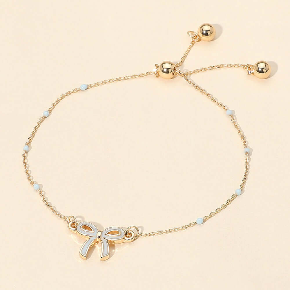 The Tied to You - Light Blue Bracelet features a gold bow charm, small white beads, adjustable clasp with two gold beads at the ends—perfect for stacking. Displayed on a light beige background.