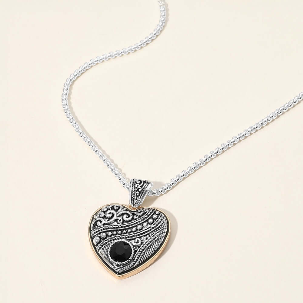The Hearts & Heritage - Black Two Tone Necklace is a vintage-inspired silver chain with a heart-shaped pendant, gold-toned border, and small black gemstone—an elegant, timeless accessory for any occasion.
