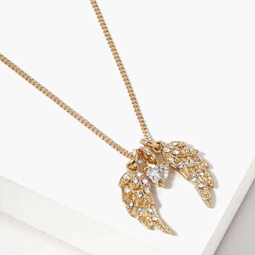 The Celestial Whispers - Gold Necklace features two crystal-encrusted angel wing pendants and a small heart-shaped crystal charm, elegantly displayed on a white surface.
