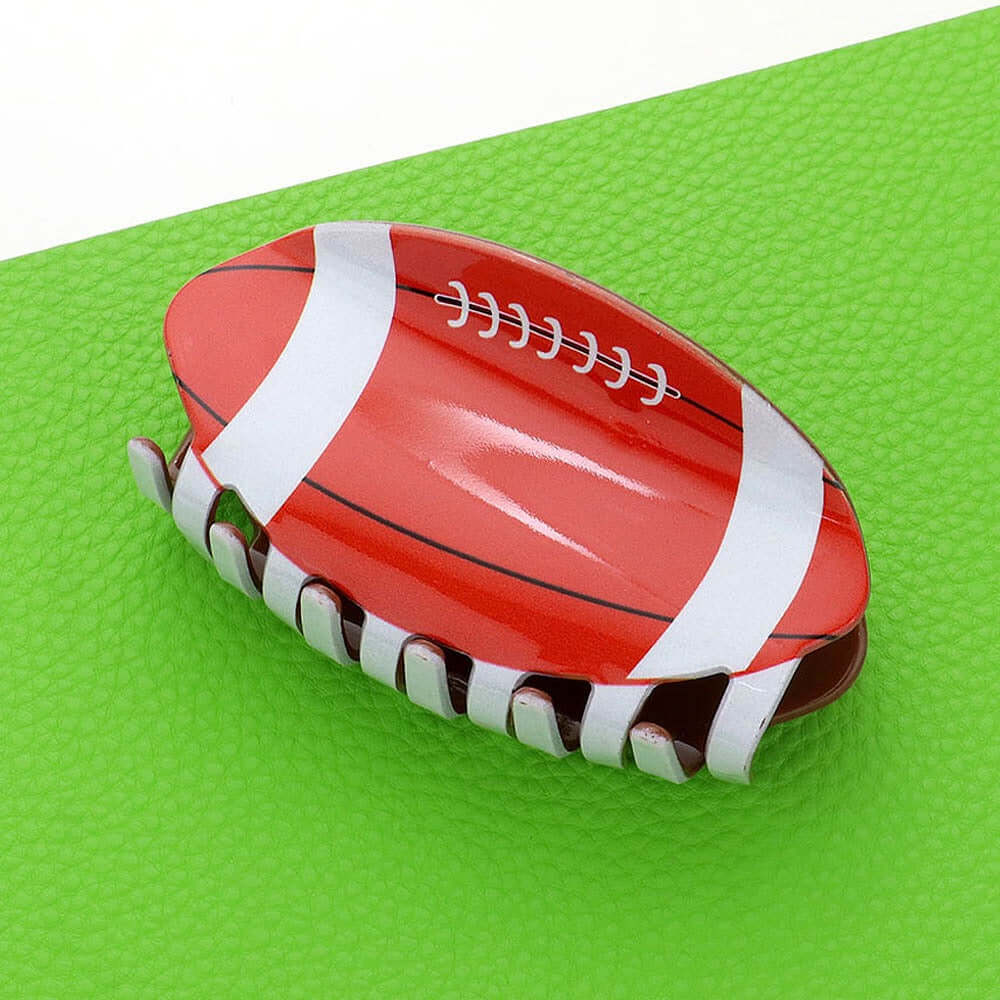 The Gridiron Grip - Brown Hairclip features an American football design with red and white details and white laces, displayed on a bright green surface—perfect for adding sporty flair to your purse hook or handbag hanger.