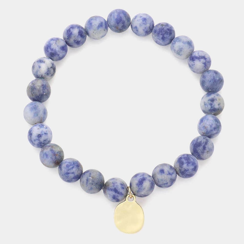 The Still Waters - Blue Bracelet features round blue and white marbled beads with a small oval gold charm, shown on a simple light background.