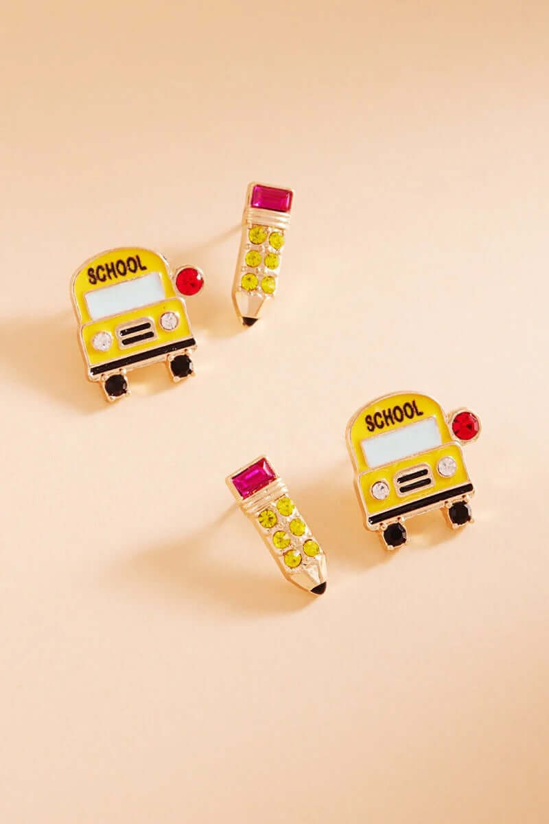 The Class Act - Multi Earring Set features mismatched back-to-school jewelry: one SCHOOL-labeled school bus earring with gem accents and a pencil stud with a pink eraser, shown on a beige background.