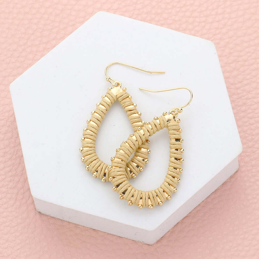 Twist & Tassel - Gold Earrings: Teardrop-shaped drops with woven detail and small gold beads, shown on a white hexagonal platform against a pale pink background—versatile boho earrings for any outfit.