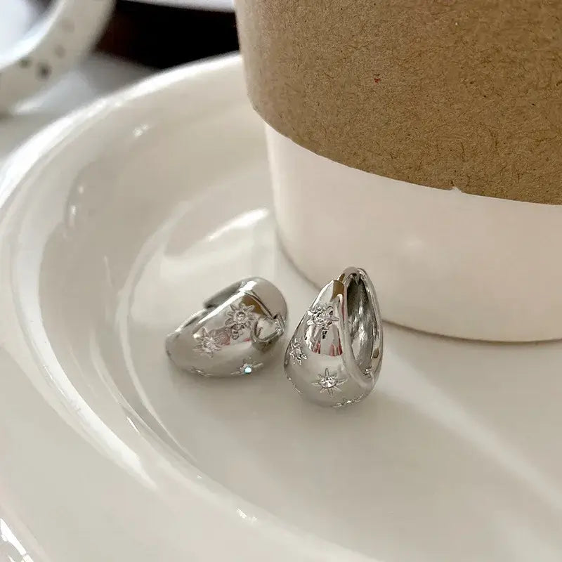 A pair of Subtle Sparkle - Silver Hoop Earrings rests on a white ceramic plate beside a brown paper cup.