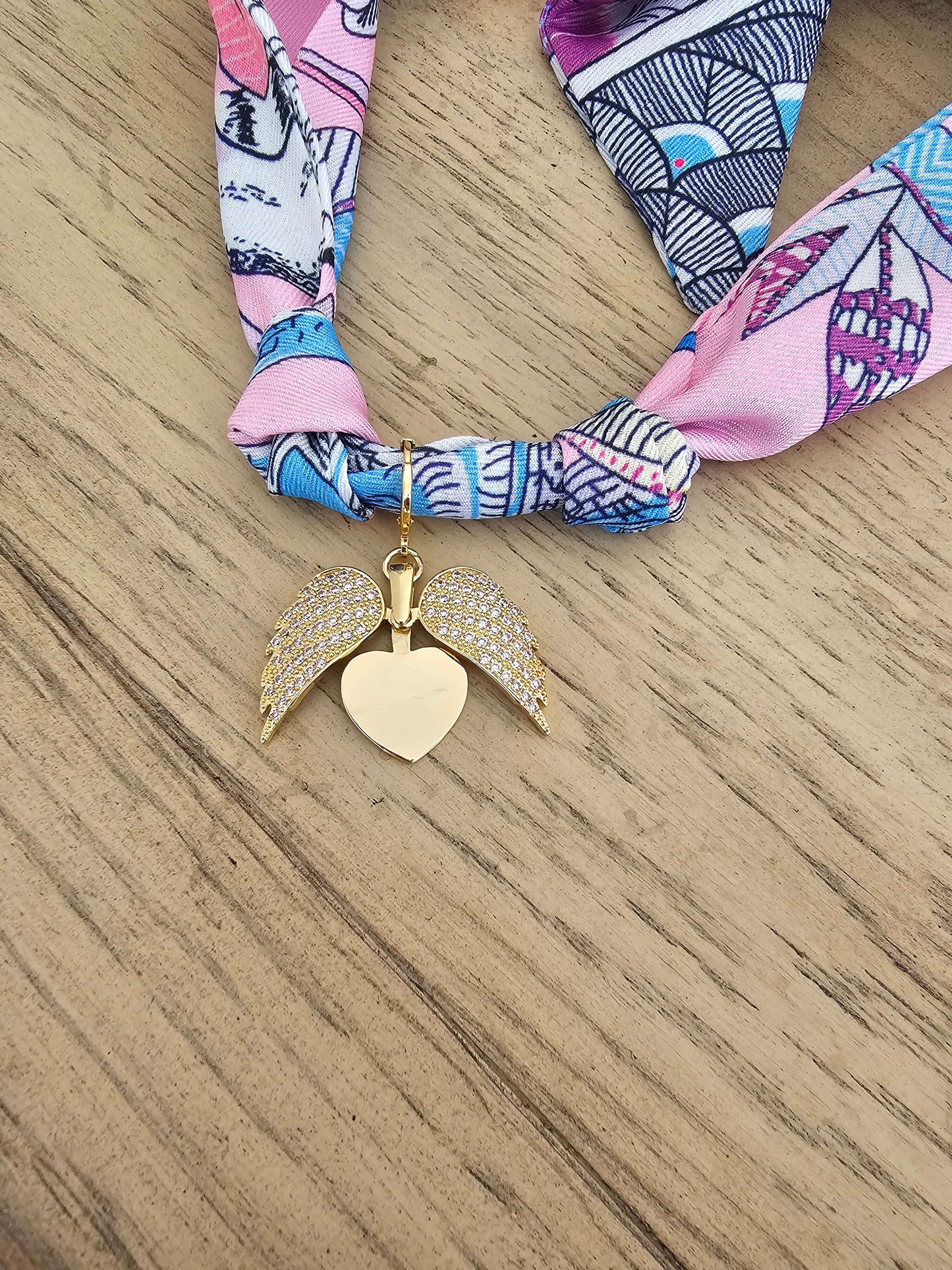 The Exclusive Pink & Purple Scarf Necklace features an 18k gold-plated angel wings charm and rests on a light wooden surface—a unique, handmade jewelry piece.
