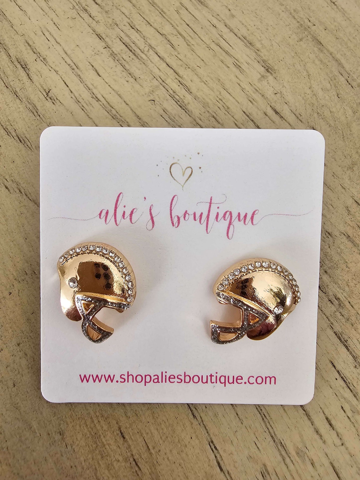The Game Face On - Multi Earrings are gold football helmet studs with rhinestone sparkle, displayed on an alies boutique card (www.shopaliesboutique.com), perfect for game-day style and shown on a light wood surface.