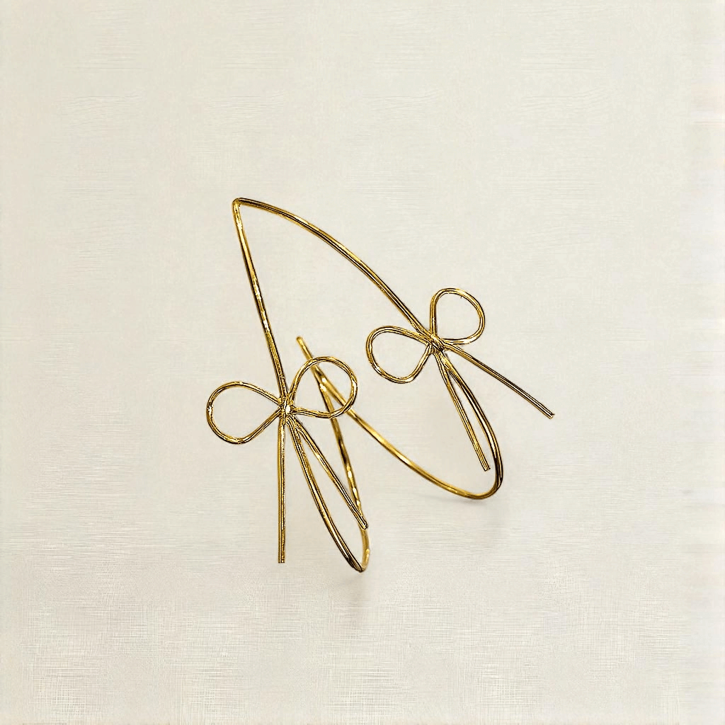The Ladylike Bows- Gold Hoop Earrings, shaped like delicate bows, add a feminine touch as they rest on a light, textured surface.
