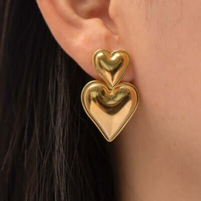 Close-up of a person wearing Double Love Gold Earrings—two stacked hearts, one smaller atop a larger—against their ear. Their dark hair is partially visible, accentuating the interlocking heart design of the earrings.