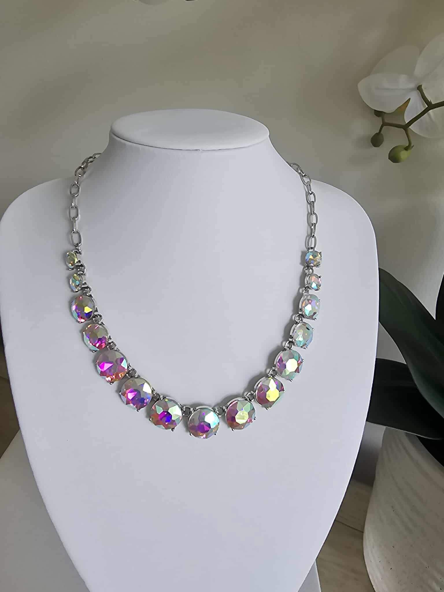 The Aurora Pop - Iridescent Necklace showcases large iridescent gems on a silver chain, displayed on a white jewelry bust with a white orchid plant in the background.