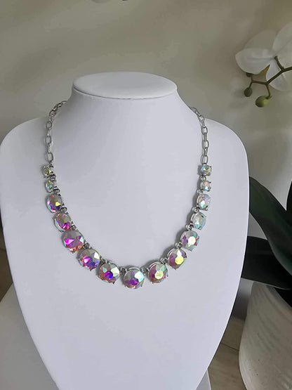 The Aurora Pop - Iridescent Necklace showcases large iridescent gems on a silver chain, displayed on a white jewelry bust with a white orchid plant in the background.