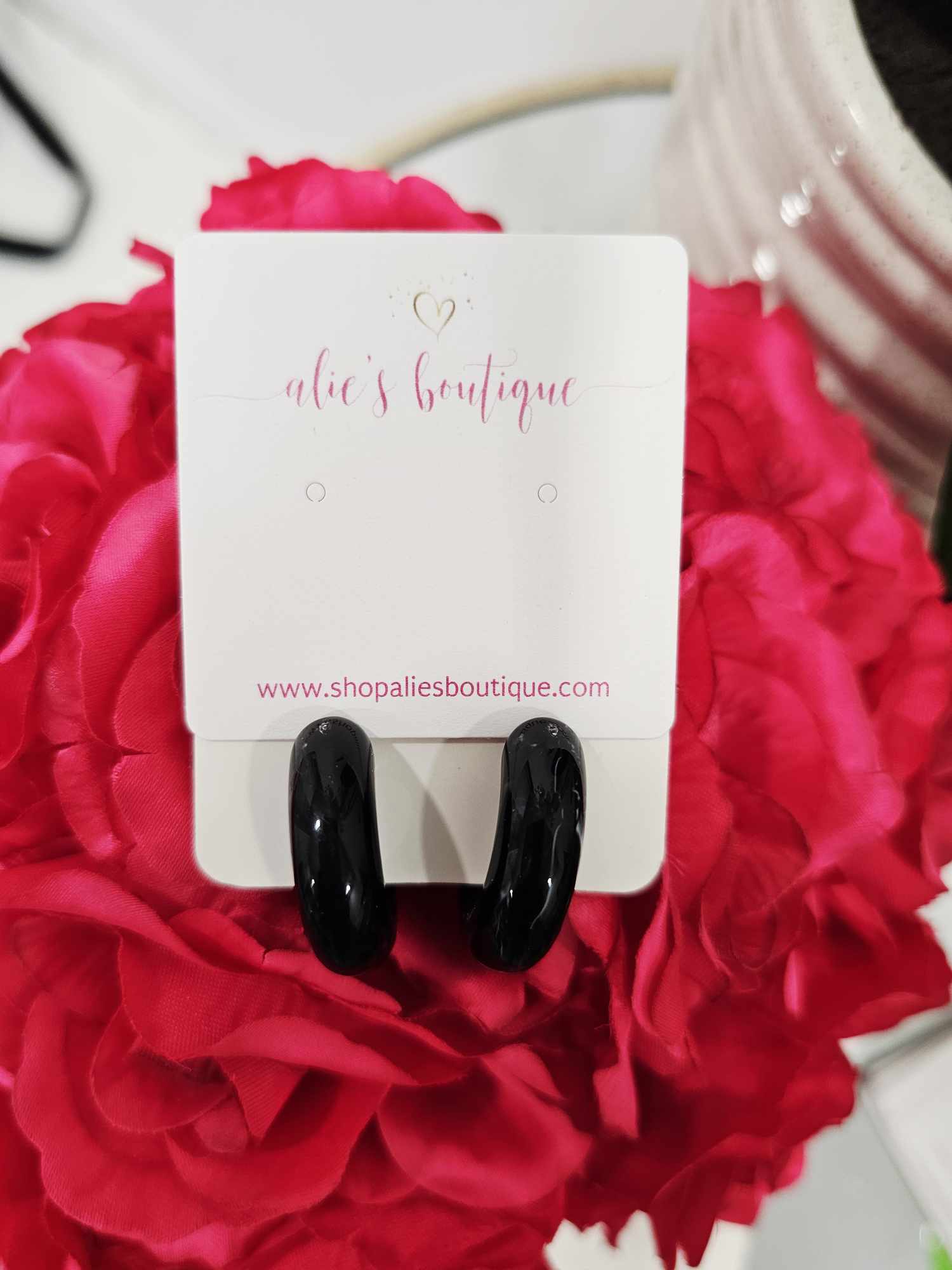 A pair of Chaotic Chic - Black Marble Acrylic Resin Hoop Earrings - WW is displayed on a white card with "alies boutique" in pink, set against vibrant pink flowers. Find these trendy earrings at shopaliesboutique.com.
