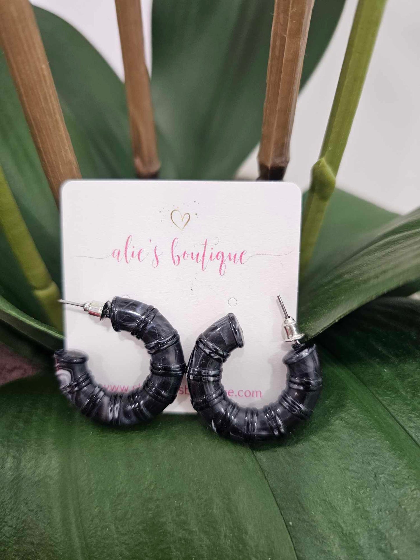 A pair of Chaotic Chic - Dark Grey Acrylic Resin Hoop Earrings - WW are displayed before an alies boutique card, set against green leaves and plant stems. These trendy hoops are perfect to elevate any look.