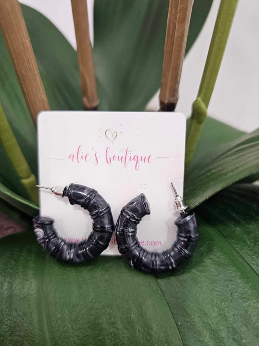 A pair of Chaotic Chic - Dark Grey Acrylic Resin Hoop Earrings - WW are displayed before an alies boutique card, set against green leaves and plant stems. These trendy hoops are perfect to elevate any look.