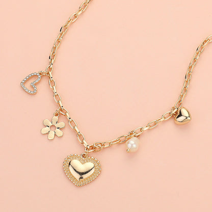 The Love Blossoms - Gold Charm Necklace features five elegant charms—a rhinestone heart, gold flower, large gold heart, pearl bead, and solid gold heart—displayed on a soft pink background.
