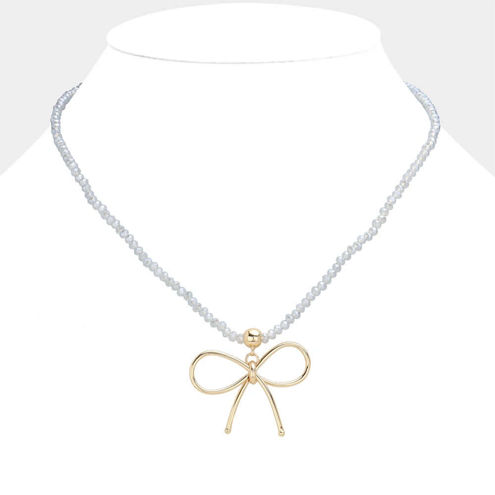 The Soft Intentions - Blue Bow Necklace features delicate beads and a gold bow pendant, perfect for layering and elegantly displayed on a white stand.