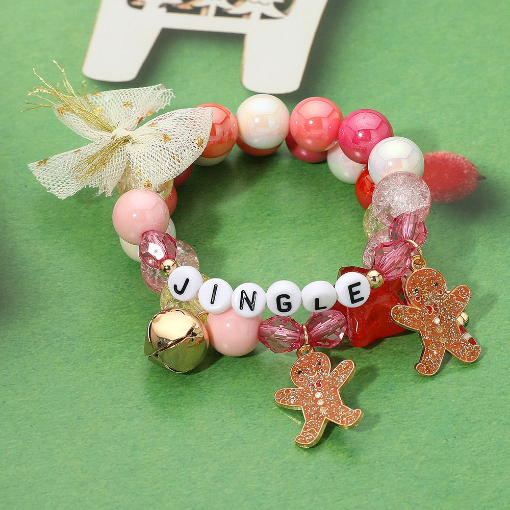 The Merry Maker - Gold Multi Bracelet features pink, red, and white beads, gingerbread charms, a gold bell, a gold bow, and “JINGLE” in white letter beads—festive jewelry shown on a green background.