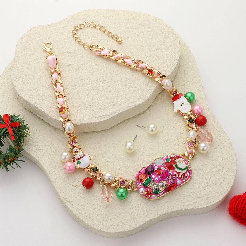 The Jingle Luxe - Holiday Glam Necklace features a colorful gold chain with pink, red, and green beads, pearl accents, festive charms—including a Christmas pendant—shown on cream platforms beside matching pearl stud earrings.