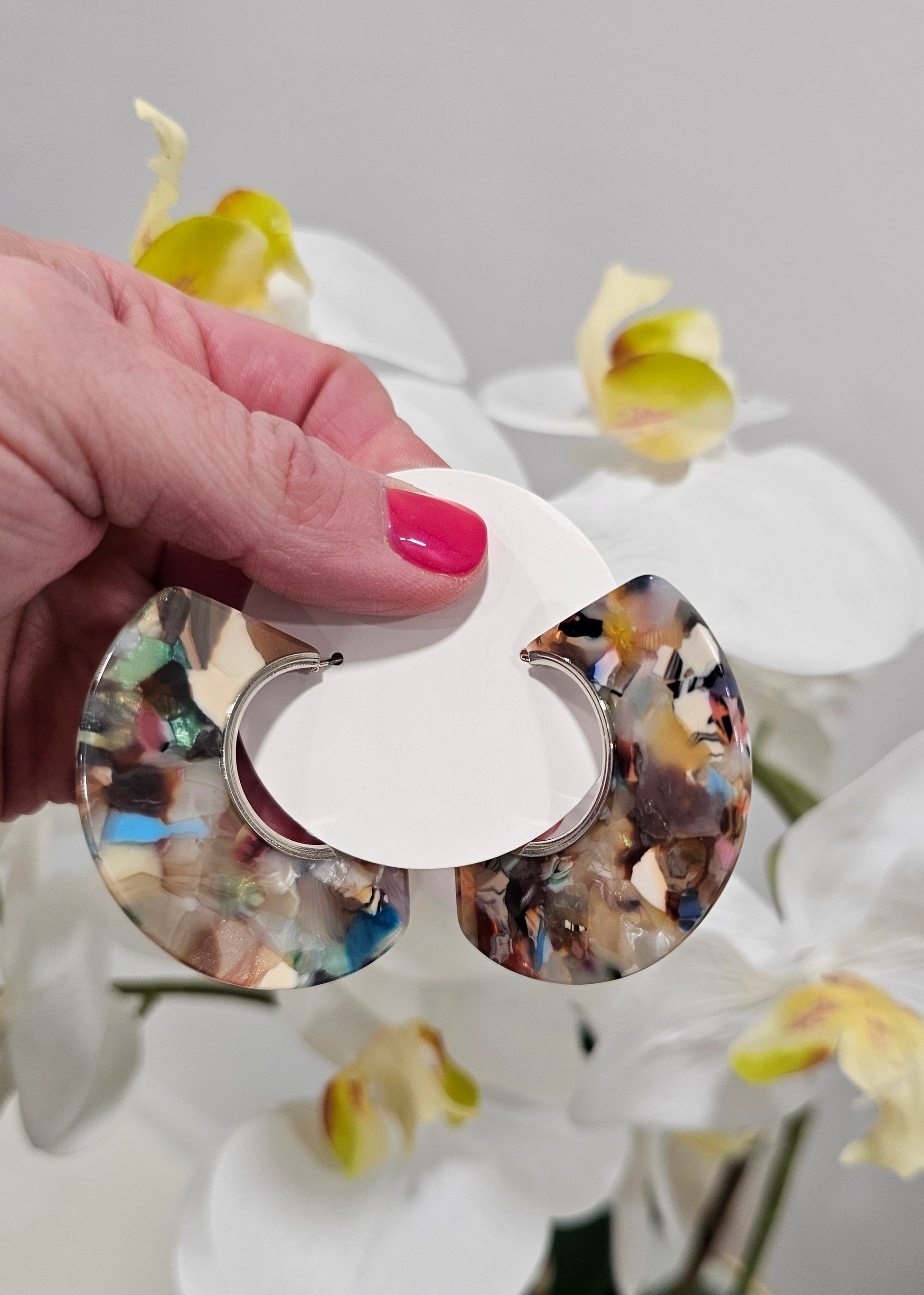 A hand with pink nail polish holds the Kaleido-hoops - Mocha Multi Earrings, chunky crescent-shaped accessories with a colorful marbled pattern, pictured against white orchids—ideal for bold jewelry lovers.